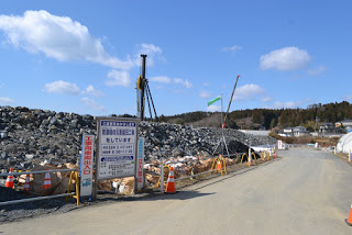 道路の左側に石のブロックが高く積まれ、複数の重機が見える工事現場があり、道沿いに工事の案内看板が立っている様子の写真