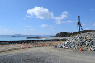遠くに山並みが見える海岸近くの道沿いに、赤白のコーンが並び、右側には大きな石のブロックの山があり、その奥に背の高い重機が見える写真