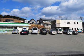 広いアスファルトの駐車場に車が横一列に停まり、その後ろにはコンビニと白い建物の仙台銀行が並んで建っている写真