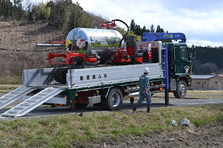 大型トラックから赤い車体に銀色のタンクがついた液肥散布車が降ろされようとしている写真