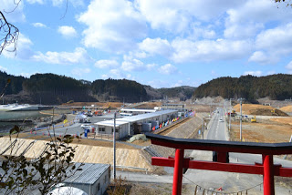 神社の赤い鳥居の先に長い一本道が続き、その右側の平らな土地に工事車両が見られ、左側のかさ上げした土地の上には小胆外の建物が並んでいる様子を高台から撮影した写真