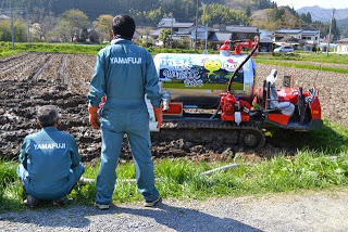 「YAMAFUJI」と背中に書かれた薄緑色の作業服を着た二人の男性が、畑に停まっている液肥専用散布車を見つめている後ろ姿の写真