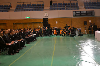 黒いスーツを着た大勢の参列者が並んだ椅子に座り、一角には楽器を手にした黒い衣装を着たオーケストラの一団が座っている写真