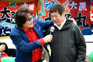 大漁旗を背景に、亜KIRAさんが一人の男性にマイクを向けてインタビューしている場面を撮影した写真