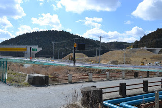 小高い山の手前に土が山のように積まれた道路工事の現場があり、その手前を通る道路の先に緑色の案内板と、建設中の橋梁が見える写真