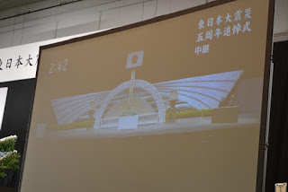 会場の大きなスクリーンに東日本大震災五周年追悼式の中継映像が映し出されている写真