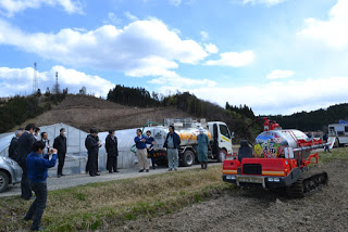 液肥散布車が停まった畑の横の道路に関係者や見学者などが大勢集まって、話をしている様子の写真
