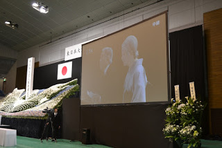 白菊の生花装飾の隣に置かれた大きなスクリーンに天皇陛下御夫妻が映し出されている写真