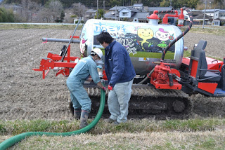 作業服を着た2人の男性が、畑に停っている液肥散布車に緑色のホースを繋げている写真