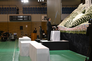 ステージ前に置かれた白いクロスが掛けられた演台に立ち、慰霊碑を見上げながら追悼の言葉を述べる男性の写真