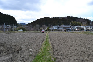 低い山や住宅を背景にした畑の真ん中に細い草道が伸び、奥に赤い液肥散布車が見える写真