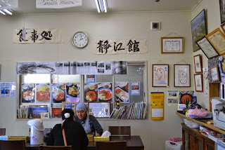 壁に「食事処 静江館」と大きく書かれた張り紙や料理の写真が多数貼られている店内で、テーブルに座った女性と齋藤さんが会話している様子の写真