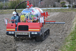 作業服を着てヘルメットをかぶった男性が運転する赤い液肥散布車のタンクから伸びたパイプに等間隔に空けた穴から液肥が散布されている様子の写真