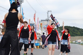 赤い法被を羽織った若い女性たちが、笑顔で右手を上げながらリズムに乗って踊っている写真