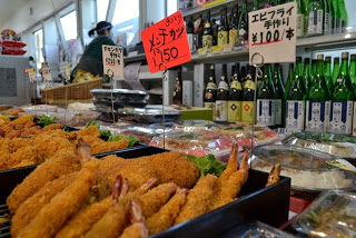 お酒の瓶が並んだ陳列棚の前のスペースに、札が立っている前に揚げたてのエビフライやメンチカツなどがトレイに盛られた惣菜売り場の写真