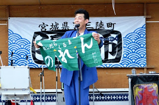 ステージ上で青いスーツ姿の男性が、緑色ののれんを掲げて観客に向かってパフォーマンスしている写真