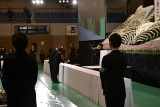 ステージ前に設置された白いクロスが掛けられた長い献花台の中央に花を供え、慰霊碑に向かって礼をしている男女の写真