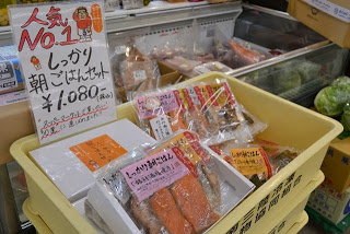 値札と「人気No.1 しっかり朝ごはんセット」と書かれた手書きのポップが立っているプラスチックの大きな黄色い箱に、パック入りの鮭や魚の切り身が並んで詰められている写真