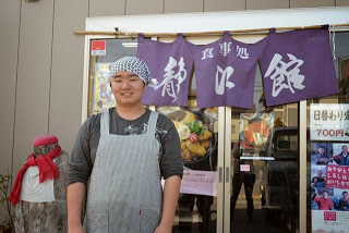 「食事処 静江館」と書かれている暖簾が設置された店の入口の前で、頭にマルニ手ぬぐい巻きしエプロンを着けた齋藤楓稀さんが笑顔で立っている写真