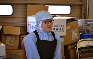 段ボール箱や備品が積まれた倉庫内で、白い帽子と黒いエプロンを着た金野幾美さんが話している写真