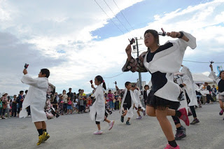 白い羽織と黒の衣装を着た子どもたちが、鳴子を持って地面を蹴るように元気に踊っている写真