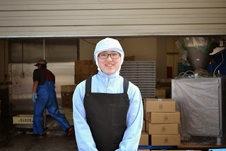 重機や段ボールが積まれて置かれた工場の前で、作業服と帽子、黒いエプロンをつけた金野幾美さんが笑顔で立っている写真