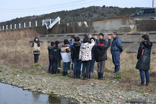 川岸で水戸辺川サケ部会の方々と向かい合って立った子どもたちのうち、数名が右手を挙げている写真