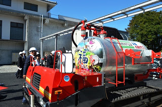 色鮮やかな絵が描かれたタンクを載せた赤い車両を関係者が屋外で見学しており、建物の壁と配管設備が背景にある写真