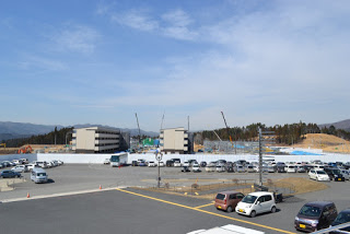 手前の広い駐車場に自動車が並び、奥には白い壁で囲まれた工事中の建物とクレーンがある復興住宅建設現場が見える写真