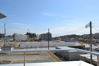 金属製の手すりの内側から撮影した、駐車スペースの先に、白い壁で囲まれた、クレーンや建設中の建物が並んでいる工事現場が見える写真