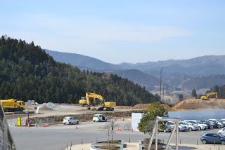 右側にたくさんの車が停まった駐車場があり、その奥で複数の黄色いショベルカーが作業をしている様子の写真