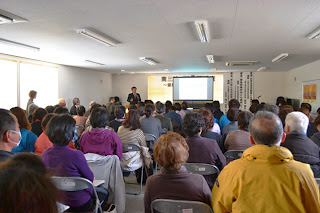 講師の男性が前方で話をしており、多数の参加者が会場の椅子に座って前を向き、熱心に講座の内容に耳を傾けている様子を写した写真