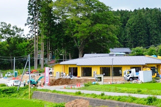 黄色い外壁の建物の周りで工事車両や作業員が作業しており、建物の背後には大きな木と森が広がっている写真