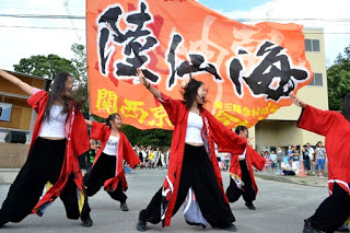 赤い法被を着た女性たちが大きな「陸仙海」と書かれたオレンジ色の旗の前で、両手を広げて力強く踊っている写真