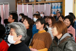 椅子に座っている多数の参加者が、前方の講演者の話を笑顔で楽しそうに聞いている様子を写した写真