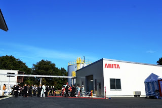 青空の下、「AMITA」と書かれた白い建物の前で関係者が集まっている写真