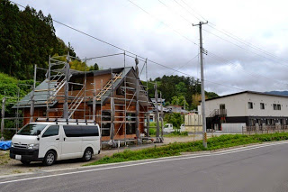 道路沿いにある建設中の木造住宅の周りに足場が組まれており、白いワゴン車が手前に停まっている写真