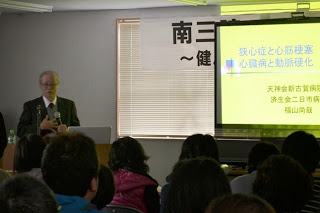 スクリーンに資料を映し、壇上でマイクをもって話をしている福山尚哉先生の写真