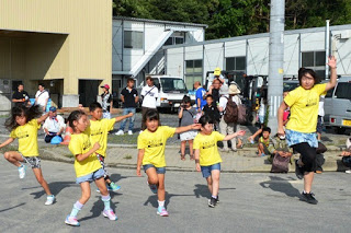 黄色いTシャツを着た子供たちと女性の指導者が、ジャンプしながら元気に踊っている写真