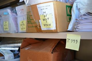 段ボール箱やケールが棚に並び、付箋で分類ラベルが貼られている書類保管エリアの写真