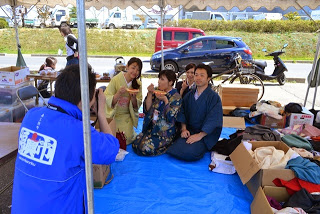 テントの下の青いシートの上で着物姿の男女が座り、カメラに向かって笑顔でポーズを取っている様子の写真