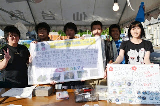 テントの下で若者たちがモアイ像について書かれた手作りのポスターを掲げ、モアイバッジの販売を行っている写真