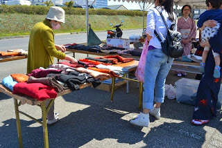 道路沿いのテントの下で女性客たちが机の上に並べられた着物や布を手に取って選んでいる様子の写真