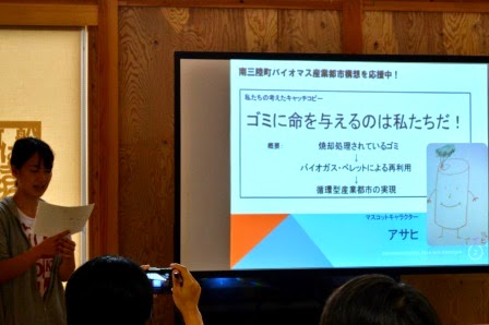 資料が映し出されたスクリーンの左側に立つ女性のグループが考えた「ゴミに命を与えるのは私たちだ！」のキャッチコピーが写され、原稿を読みながらゴミの有効活用による持続可能な社会を訴えた発表を行っている写真