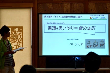 緑のシャツを着た男性のグループが考えた「循環＋思いやり＝鏡の法則」のキャッチコピーが写され、スライドを見ながらバイオマス活用の理念を伝える活動の発表を行っている写真