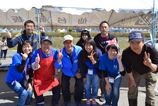 屋外のテント前で青いスタッフTシャツを着た人たちが2列に並び笑顔でピースサインをしている写真
