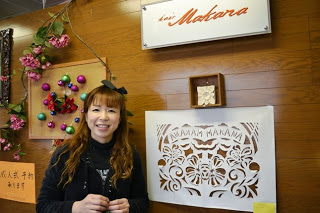 木目の壁に白い切り絵作品が飾られ、その横で笑顔の女性が立っている店内の一角で写した写真