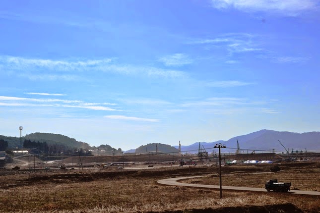 広々とした造成中の土地が広がり、奥には山と重機が見え、手前には未舗装の道を走る車が一台写っている、工事現場の写真
