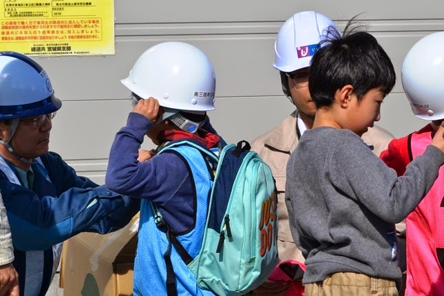 リュックを背負った子どもが青いベストを着用し大人にヘルメットを調整してもらっており、周囲には他の子どもたちもヘルメットをかぶっている様子が見られる、出発前の支度の写真。