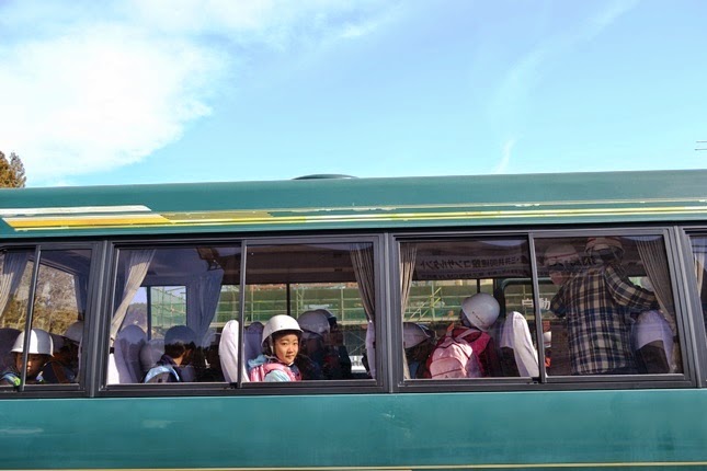 子供たちがヘルメットをかぶってバスの中に座っており、そのうちの一人が窓越しにこちらを見ている様子の写真